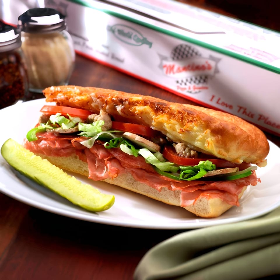 Mancino's grinder sandwich with meats, cheese, lettuce, tomatoes, and a pickle spear on a white plate.